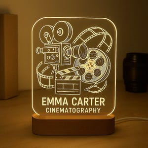 May include: A film-themed night light with a wooden base. The acrylic design features a vintage film camera, film reel, and clapperboard. The text "EMMA CARTER CINEMATOGRAPHY" is displayed on the front.