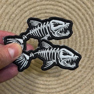 Puede incluir: Dos parches bordados con un diseño de esqueleto de pez. Los parches tienen un borde negro con costuras blancas que delinean los huesos, la cabeza y la cola del pez. Los parches están en la mano de una persona.