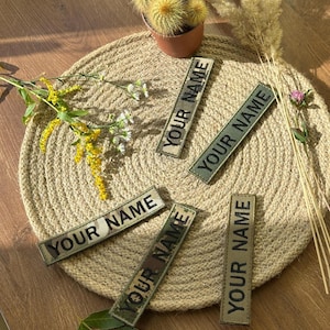 Pode incluir: Cinco patches retangulares com o texto "YOUR NAME" em letras pretas. Os patches t&ecirc;m um padr&atilde;o de camuflagem e est&atilde;o dispostos sobre um descanso de panela de juta tecida. Um pequeno cacto em vaso e flores secas tamb&eacute;m est&atilde;o presentes.