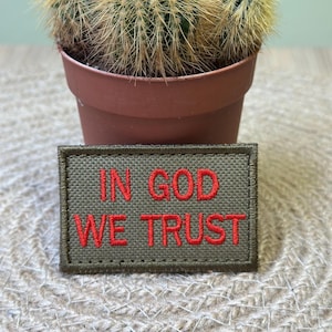 May include: An olive green patch with the embroidered text "IN GOD WE TRUST" in red. The patch is rectangular and has a stitched border. A small potted cactus is in the background.