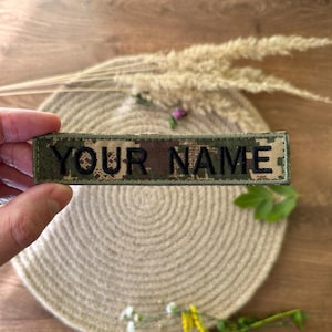 Pode incluir: Um patch retangular com o texto "YOUR NAME" em letras pretas. O patch tem um padr&atilde;o de camuflagem em tons de verde, castanho e bege. O patch &eacute; exibido em um fundo neutro.