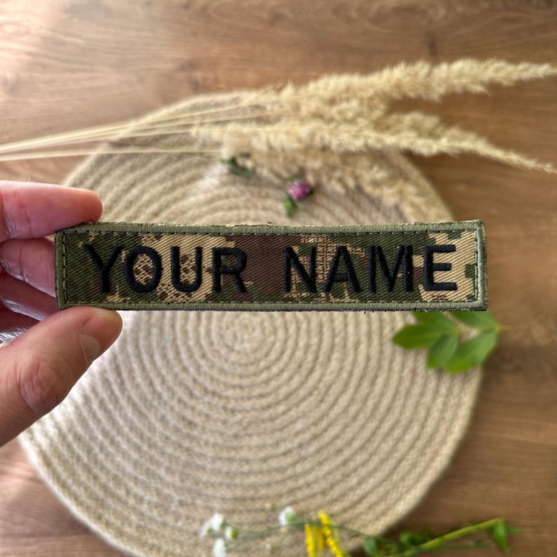 Pode incluir: Um patch retangular com o texto "YOUR NAME" em letras pretas. O patch tem um padr&atilde;o de camuflagem em tons de verde, castanho e bege. O patch &eacute; exibido em um fundo neutro.