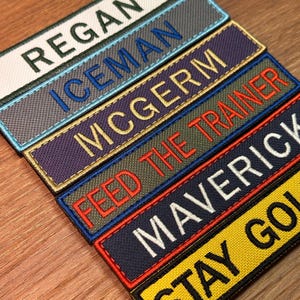 May include: Close-up of several embroidered name patches in various colors. The patches display names and phrases in white, blue, and gold thread. The patches are rectangular and have a textured appearance.