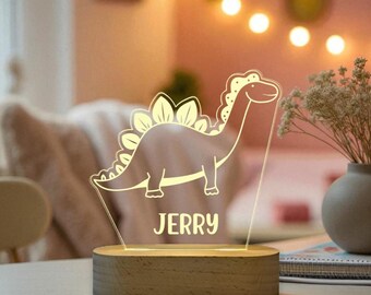 Puede incluir: Una luz nocturna LED con forma de dinosaurio con el nombre "JERRY" iluminado en blanco. El dinosaurio está delineado en un cálido brillo amarillo, colocado sobre una base de madera. El fondo está suavemente iluminado con luces de cadena.