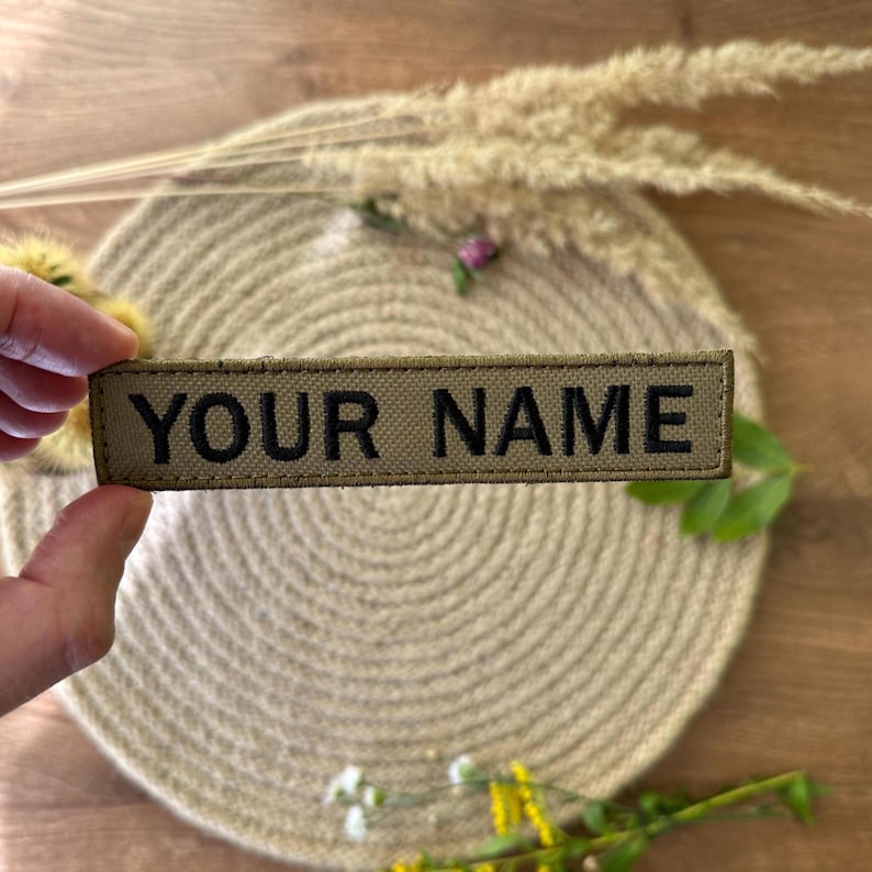 Pode incluir: Um patch retangular verde-oliva com o texto "YOUR NAME" em letras pretas. O patch &eacute; segurado por uma m&atilde;o, com um fundo neutro de um tapete tecido e plantas secas.
