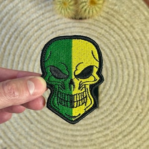 Pode incluir: Um patch de caveira verde e amarelo com contorno preto. A caveira é dividida verticalmente, com o lado esquerdo verde e o lado direito amarelo. O patch é segurado por uma mão contra um fundo neutro.