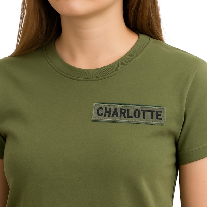 Pode incluir: T-shirt verde-oliva com gola redonda. Um patch retangular com o nome "CHARLOTTE" em letras pretas &eacute; costurado na camisa. A camisa &eacute; feita de um material macio e confort&aacute;vel.