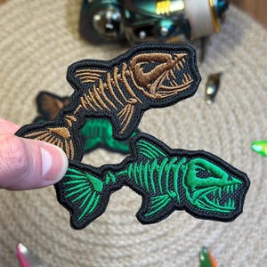 May include: Embroidered patches with fish skeleton designs. One patch has a brown skeleton on a black background, and the other has a green skeleton on a black background. The patches are held up against a neutral background.