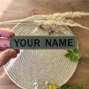 Pode incluir: Um patch retangular verde-oliva com o texto "YOUR NAME" bordado em preto. O patch &eacute; segurado por uma m&atilde;o, com um fundo neutro de um tapete tecido e plantas secas.