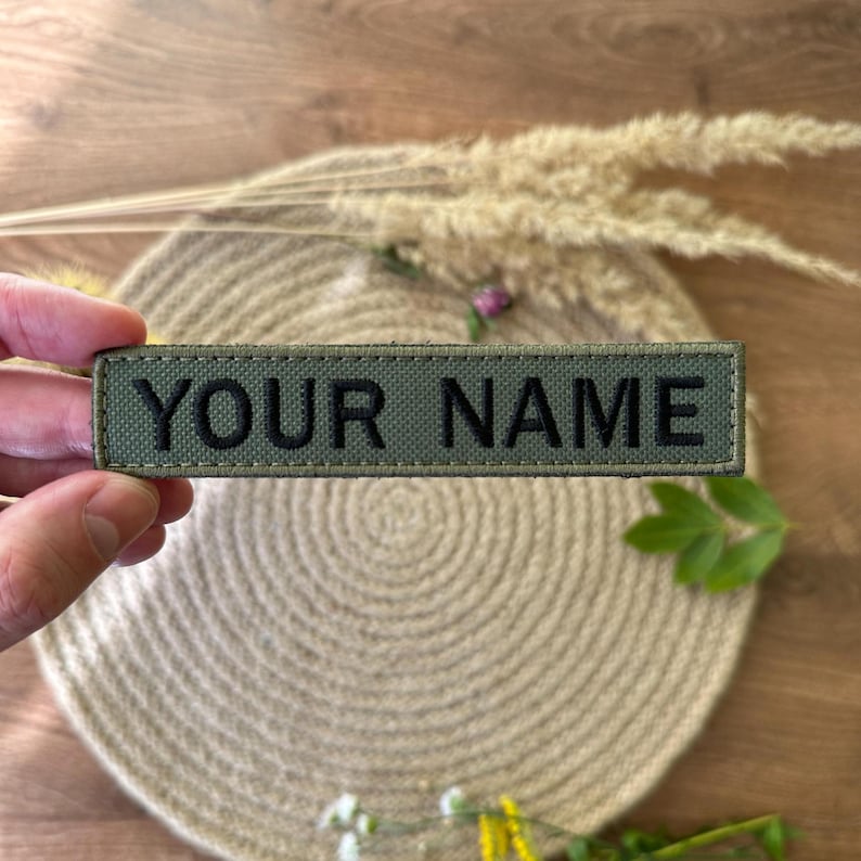 Pode incluir: Um patch retangular verde-oliva com o texto "YOUR NAME" bordado em preto. O patch &eacute; segurado por uma m&atilde;o, com um fundo neutro de um tapete tecido e plantas secas.