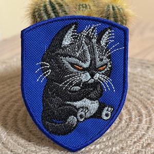 Può includere: Toppa ricamata raffigurante un gatto nero scontroso con occhi arancioni e baffi bianchi, su sfondo blu a forma di scudo. Il gatto è seduto con le zampe incrociate. Per abbigliamento o accessori.