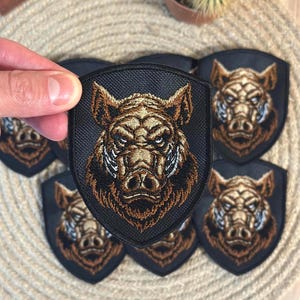 Könnte beinhalten: Gestickte Aufnäher mit einem detaillierten Wildschweinkopf-Design. Die Aufnäher haben eine schwarze Schildform mit einem braunen und goldenen Wildschweinkopf. Das Wildschwein hat einen wilden Ausdruck und weiße Akzente. Geeignet für Jacken, Taschen oder andere Stoffartikel.