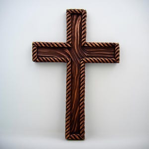 以下が含まれることがあります： 濃い茶色の仕上げとロープのような縁取りが施された木製の十字架。十字架には木目の詳細な模様があり、白い背景に表示されています。十字架は信仰と精神性の象徴です。