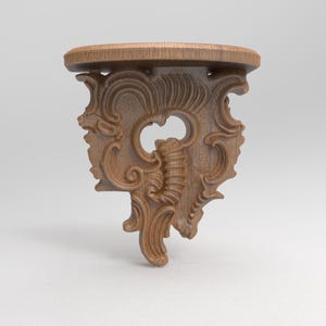 Puede incluir: Estante de pared de madera con un diseño tallado ornamentado. El estante tiene una parte superior redonda y un soporte decorativo con patrones arremolinados y un elemento central estilizado. La madera es de color marrón claro natural.