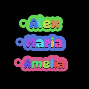 May include: Three colorful name tags with the names "Alex", "Maria", and "Amelia" in bubble letters. Each name is on a different colored background: green, blue, and pink, respectively. The tags have a small hole at the top.