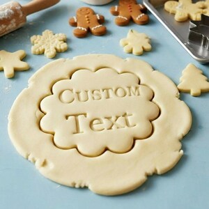 May include: A close-up shot of cookie dough with the words "Custom Text" imprinted. Several baked cookies in various shapes, including gingerbread men, snowflakes, and trees, are scattered around. A rolling pin is visible in the background.
