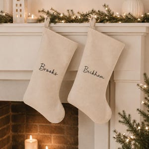 May include: Two beige fabric Christmas stockings hanging side by side. The left stocking features the name Brooks embroidered in black script, and the right stocking features the name Beckham embroidered in black script, both with hanging loops.