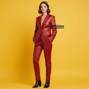 Op de afbeelding: Een rood leren pak, bestaande uit een blazer en broek, wordt getoond tegen een gele achtergrond. De blazer heeft een sluiting met &eacute;&eacute;n knoop en de broek is op maat gemaakt. De outfit wordt gedragen met zwarte enkellaarsjes. De tekst "Leather Synergy" is zichtbaar.