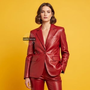 Op de afbeelding: Een rode leren blazer en bijpassende broek. De blazer heeft een sluiting met &eacute;&eacute;n knoop en twee voorzakken. De outfit is een diepe tint rood, tegen een gele achtergrond. Het logo "Leather Synergy" is zichtbaar.