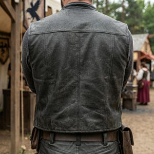 May include: A weathered, dark gray leather vest with a classic design. The vest features a tailored fit, visible stitching, and a slightly distressed appearance. The person is wearing a gray sweater underneath. A brown leather belt and matching pants are also visible.