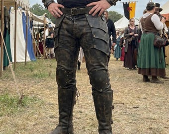 Op de afbeelding: Bruine leren broek met gelaagde pantserachtige details, kniebeschermers en bijpassende laarzen. De broek heeft een gevlekt, verouderd uiterlijk en is vastgemaakt met een riem. De laarzen zijn kniehoog met gespen en veters.