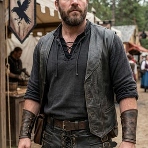 May include: A dark gray long-sleeved shirt with a laced neckline, worn under a black leather vest. The outfit includes a brown leather belt, matching leather cuffs, and a leather pouch. The background shows a medieval market setting.
