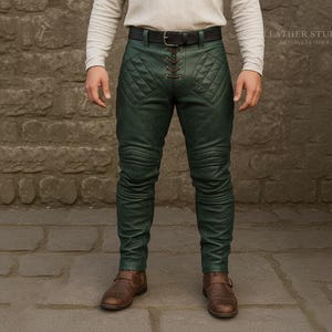 Puede incluir: Pantalones de cuero verde con detalles acolchados en los muslos y las rodillas. Los pantalones tienen un cierre de cordones en la cintura y un cinturón negro. Se usan con botas de cuero marrones.