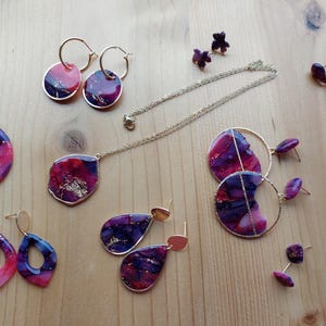 Könnte beinhalten: Eine Auswahl handgefertigter Schmuckstücke, darunter Ohrringe, Halsketten und Ohrstecker. Der Schmuck weist ein marmoriertes Design in Rosa-, Lila- und Goldtönen mit goldfarbenen Metallakzenten auf. Die Ohrringe sind in verschiedenen Formen erhältlich.