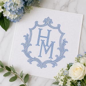 May include: A square white needlepoint canvas featuring a centered blue embroidered monogram of the letters H and M, enclosed within an ornate blue scrollwork frame. The design is stitched with a grid-like texture on a clean white fabric surface.