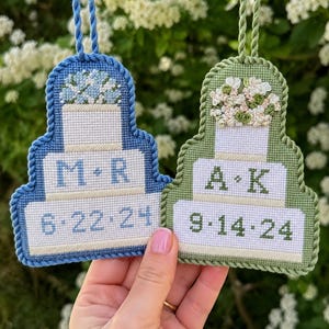 May include: Two tiered cake-shaped needlepoint ornaments with twisted cord trim and hanging loops. The blue ornament features the text M R and 6-22-24. The green ornament features the text A K and 9-14-24. Both have floral toppers.
