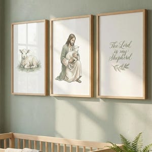 Pode incluir: Três impressões em aquarela emolduradas com molduras de madeira. As impressões retratam um cordeiro, uma figura segurando um cordeiro e o texto "The Lord is my Shepherd". As impressões são exibidas acima de um berço de madeira e uma samambaia em vaso.