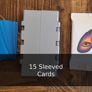 Könnte beinhalten: Kartenspielzubehör, darunter blaue Kartenhüllen mit handschriftlichem Text, eine graue Kartenaufbewahrungsbox und ein weißes Päckchen mit einem farbenfrohen Augendesign. Der Text auf der Box lautet "15 Sleeved Cards."