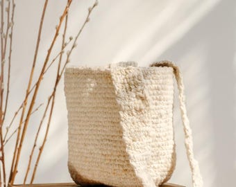 Puede incluir: Un bolso tipo cubo de color crema, tejido a mano, con una correa larga. El bolso es cilíndrico con una superficie texturizada y está sobre una caja de madera. El fondo presenta ramas desnudas y sombras suaves.