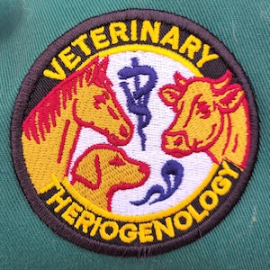 May include: Embroidered patch with the words "VETERINARY THERIOGENOLOGY" in yellow lettering. The patch features a horse, dog, and cow in yellow and red, with a blue medical symbol in the center.
