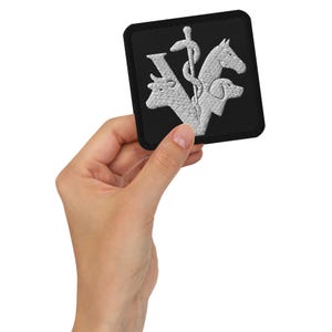 Könnte beinhalten: Schwarzer gestickter Aufnäher mit einem weißen Veterinär-Symbol, einer Schlange, die sich um einen Stab windet, und den Buchstaben "V" und "M". Das Symbol ist von den Köpfen eines Pferdes, einer Kuh und eines Hundes umgeben.