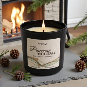 Könnte beinhalten: Eine brennende schwarze Kerze in einem Glasbehälter mit dem Text "Pinecone Nectar" auf einem Etikett. Die Kerze ist von Tannenzapfen und Grün umgeben, mit einem Kamin im Hintergrund. Die Kerze ist handgegossen und besteht aus 100 % Sojawachs.