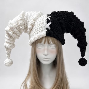 May include: A two-toned jester hat featuring a split design with one side in white and the other in black. The hat is crafted from textured crochet yarn, includes two long, curved points ending in small pom-poms, and has white ribbon lacing.