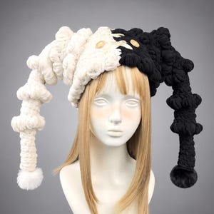 May include: A two-toned crochet hat featuring a split design with one side in white and the other in black. The hat has long, dangling, segmented horn-like extensions on each side, each finished with a pom-pom, and two small wooden buttons.