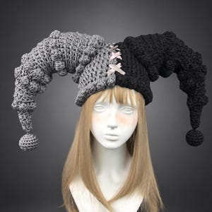 Puede incluir: Un gorro de bufón bicolor con un diseño dividido en gris y negro. El gorro de ganchillo tiene cuernos largos, curvos y texturizados a cada lado que terminan en pequeñas esferas, con una cinta rosa claro entrelazada en la parte central frontal.