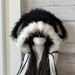Può includere: Un cappello in finta pelliccia bianca e nera con lunghe extension di capelli scuri. Il cappello presenta grandi orecchie soffici e un bordo bianco. Nastri neri e pompon aggiungono dettagli. Il cappello è esposto su una testa di manichino.