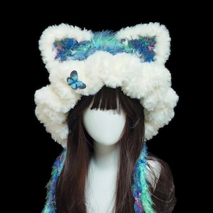 May include: A whimsical white faux fur hat with a large bow-like top, accented with blue and green fabric. A blue butterfly adorns the front. A matching scarf with blue, green, and pink accents completes the look. This unique hat is perfect for cosplay or themed events.