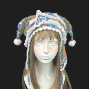 May include: A multi-colored fuzzy knit hat featuring blue, green, and orange yarn. The hat has two long ear flaps with white star-shaped pom-poms at the ends, a white lace trim along the edge, and a white ribbon bow at the top center.