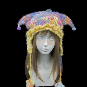 May include: A fuzzy, multi-colored jester-style hat featuring a yellow ruffled brim, two pointed side flaps with small purple and blue pom-poms at the tips, and long braided ties ending in star-shaped accents, topped with a light blue ribbon bow.