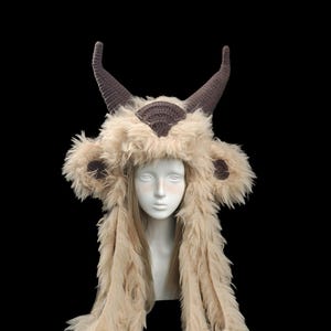 May include: A beige faux fur hat featuring two dark brown crocheted horns on top, rounded ears with dark brown centers, and two long, shaggy side panels that hang down. The hat is displayed on a white mannequin head against a black background.