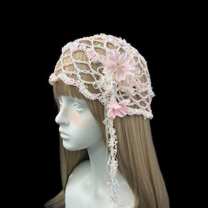 May include: A light pink crochet head covering featuring an open net pattern with iridescent sequins woven throughout. The piece is adorned with two soft pink fabric flowers on the side, with delicate beaded strands hanging down from the floral accents.