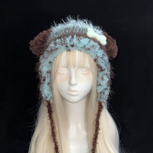 May include: A fuzzy, light blue and brown animal-ear hat featuring a white bone-shaped accent on the side. The hat has long, dangling brown braided ties that end in large, fluffy, light blue pom-poms, creating a textured, layered appearance.