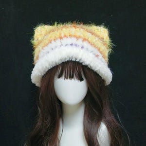 May include: A handmade cat ear hat with a fluffy, textured design. The hat features a mix of yellow, orange, and white yarn, with a white fluffy trim. The hat is displayed on a mannequin head with long brown hair.