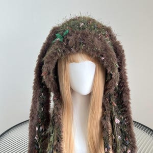 May include: A brown faux fur bunny hat with long, floppy ears. The hat is adorned with green and pink floral accents and a small green bow. The hat is designed to be worn on the head and has long, dangling ear-like extensions.
