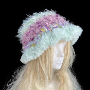 May include: A fluffy, pastel-colored hat with a wide brim. The hat features a mix of light blue, lavender, and pink yarn, creating a soft, textured appearance. The hat is displayed on a mannequin head with long, blonde hair, against a black background.