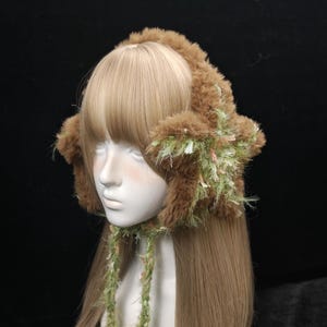 Op de afbeelding: Een bruine imitatiebont hoed met oorflappen en een bijpassende sjaal. De hoed is versierd met groene en roze accenten en bruine pompons. De hoed wordt getoond op een mannequinhoofd met lang, lichtbruin haar.